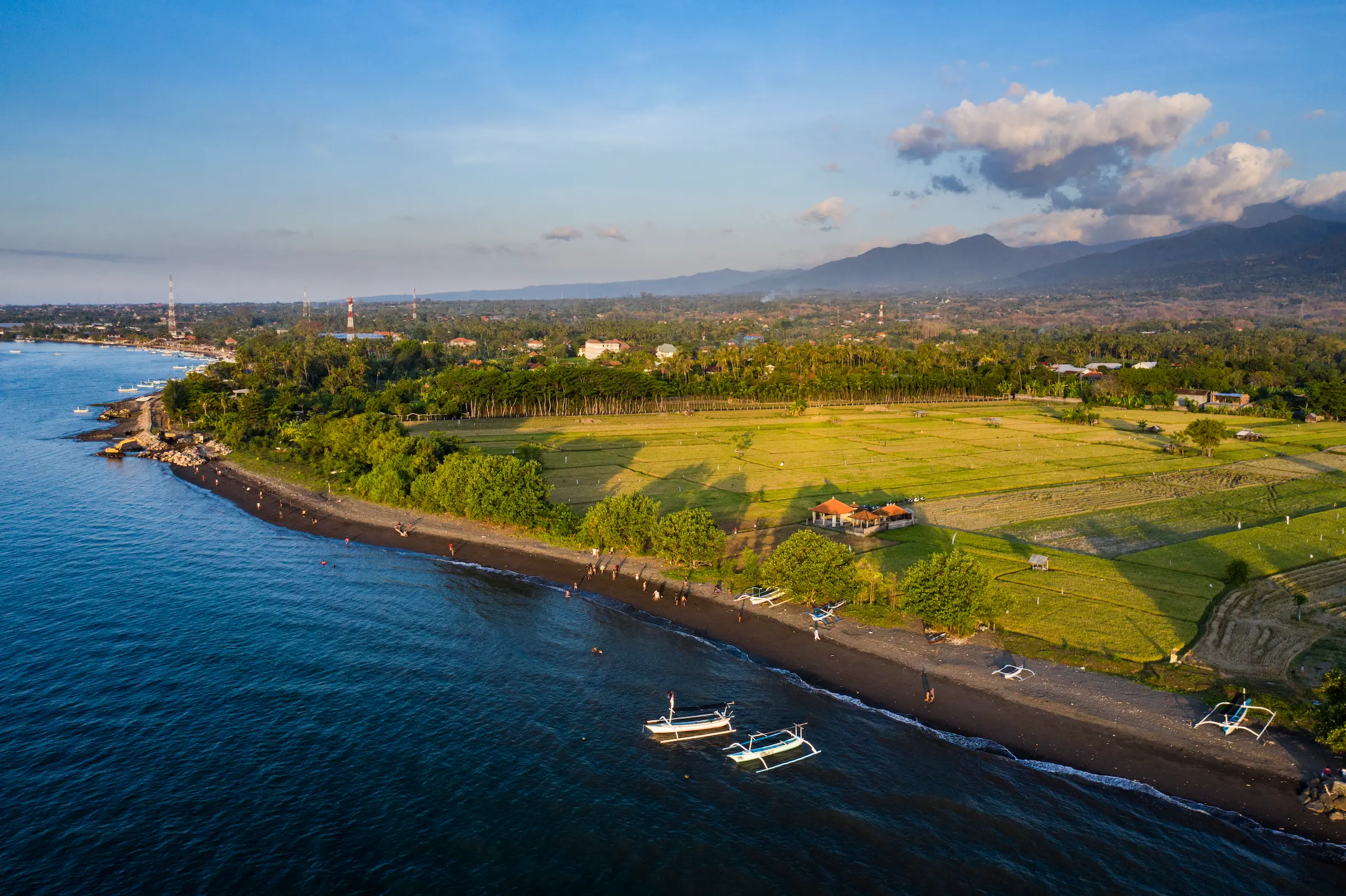 Aerial view of the North Bali coastline at golden hour — black sand beach with traditional fishing boats, rice paddies, and the mountains of Buleleng rising inland.
