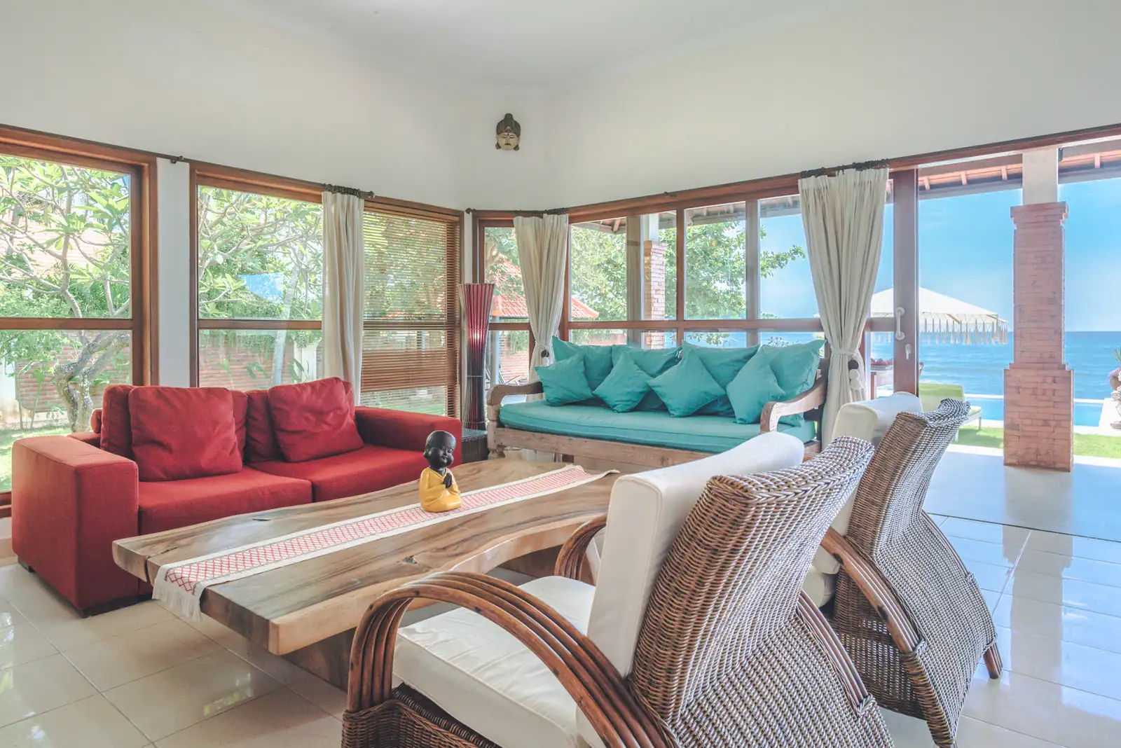 Wide living room at Primavera Beach House with red sofa, turquoise cushions, rattan chairs, and direct sea view through large windows.