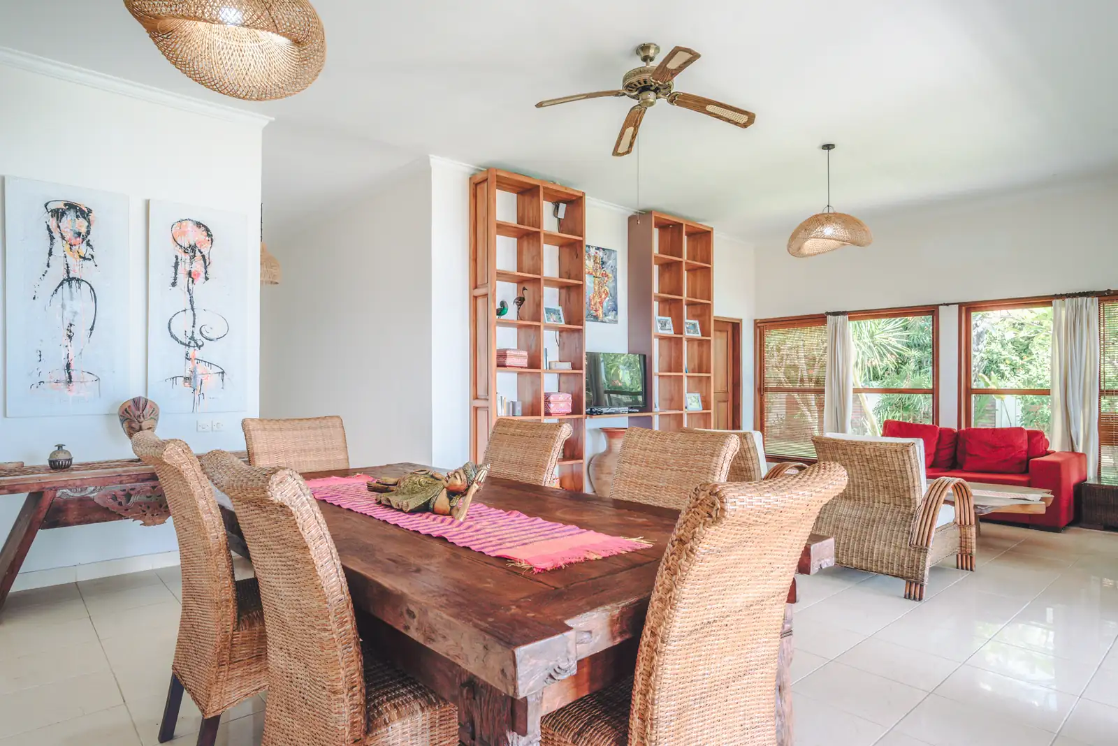 Dining area with wooden table, rattan chairs, bookshelf, and artwork at Primavera Beach House.