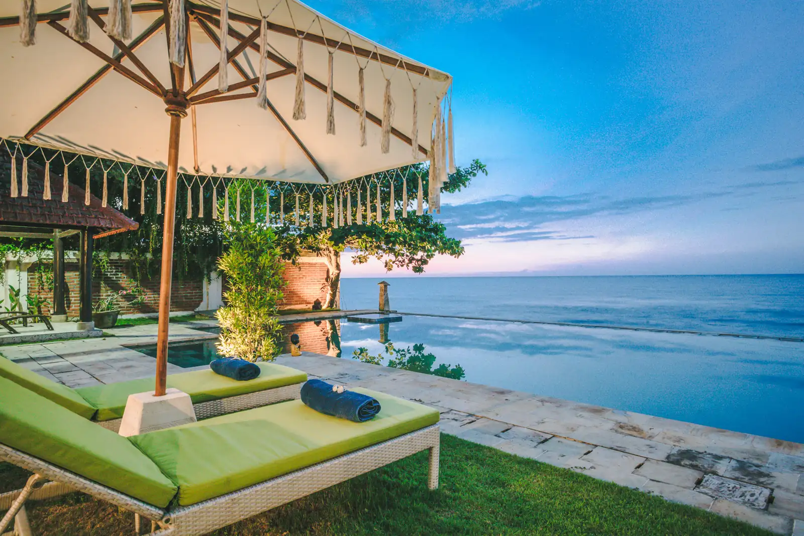 Infinity pool at Primavera Beach House at blue hour, with fringed parasol, green loungers, and the Bali Sea beyond.